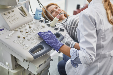 Gynecologist in white lab coat and sterile gloves using ultrasound scanner during medical examination of her patient. Smiling red-haired woman lying on daybed on blurred backgroundの写真素材
