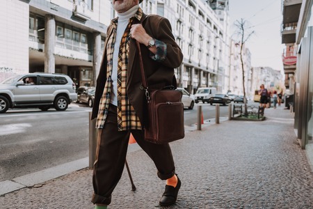 Pensioner walking in the city and holding wooden stick and briefcase with house on backgroundの写真素材