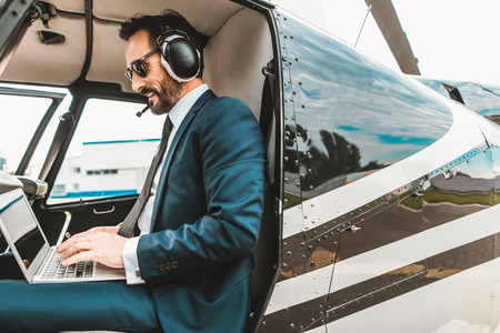 Confident elegant businessman sitting in the helicopter cabin and smiling while using his modern laptopの写真素材