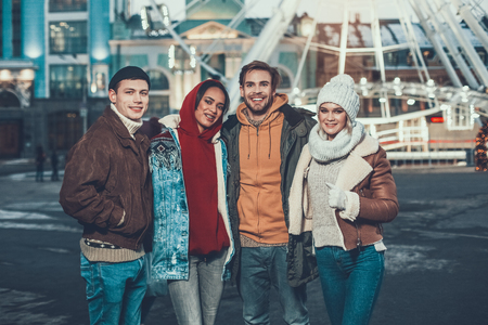 Portrait of satisfied males and cheerful women embracing together while standing outside during winter eveningの写真素材