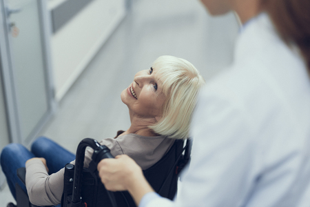 Top view with focus on delighted senior woman sitting in wheelchair. She is looking up at nurse standing behind and propelling vehicleの写真素材