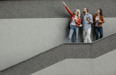 Interested young woman holding smartphone and pointing up. Others looking there with surprise. Copy space on left sideの写真素材