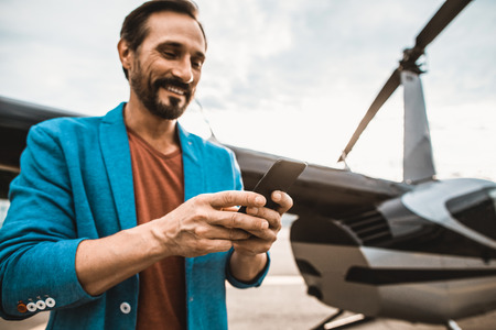 Emotional young man standing outdoors near the helicopter and smiling while looking at the screen of his modern smartphoneの写真素材