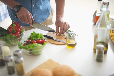 Close up of male hands with knife cutting clove of garlic. Glass of olive oil and bowls with vegetables on white kitchen tableの写真素材