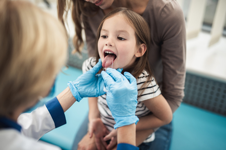 Concept of professional consultation and examination. Close up portrait of little girl with opening mouth being examined by pediatrician woman in medical officeの写真素材