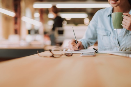 Cropped portrait of charming girl holding cup of coffee and making notes while sitting at office desk. Focus on woman handの写真素材