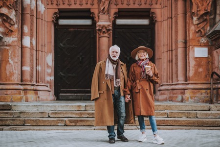 Full length portrait of stylish bearded man and his wife standing near old building. Lady in hat...