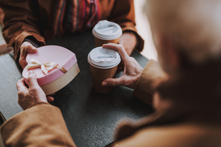 Close up of heart-shaped gift box with bow in hands of senior couple. Pensioners sitting at the table with hot drinksの写真素材