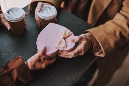 Close up of heart-shaped gift box with bow in hands of senior couple. Pensioners sitting at the...