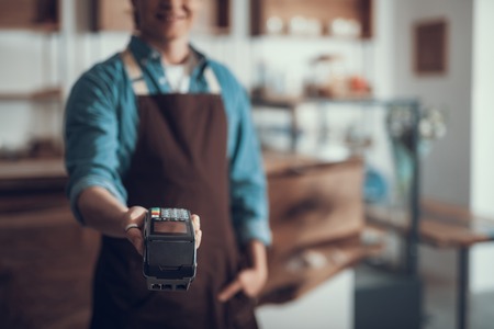 Young professional waiter standing in the cafe and putting his hand forward while holding card payment machineの写真素材