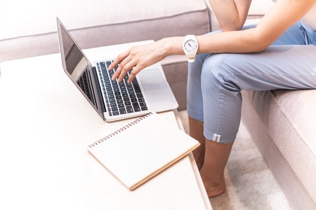 Focus on hand on woman typing on laptop keyboard. She sitting on sofa and device is on small table...