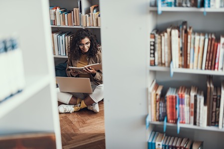 Emotional interested young lady sitting on the floor with her legs crossed and smiling while readingの写真素材