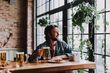 Joyful bearded gentleman with alcoholic drink sitting at the table and looking out the window with...