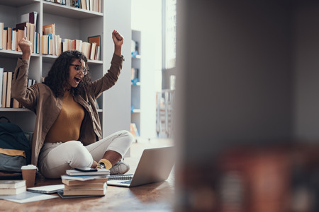 Emotional student celebrating her successful work and putting hands up while looking at the screen of modern laptopの写真素材