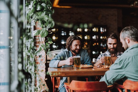 Young bearded men sitting at the table and holding mugs of beer. They looking down and smilingの写真素材