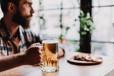 Close up of bearded gentleman holding mug of lager beer while sitting at the table. Focus on male hand with beverageの写真素材