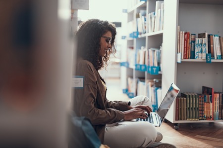 Cheerful enthusiastic young lady leaning against the bookcase and smiling while using modern laptop standing on her legsの写真素材