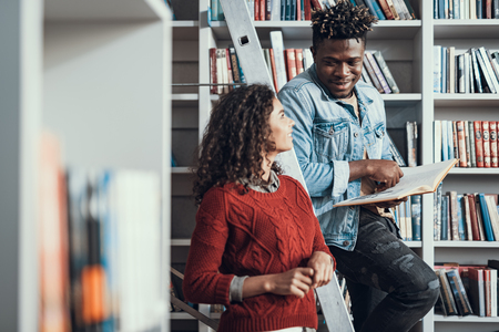 Cheerful Afro-American student being in the library with his pretty curly girlfriend and smiling to her while sitting on the step ladderの写真素材