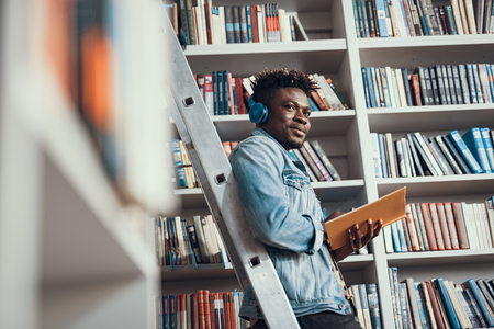 Young Afro-American student looking relaxed and smiling while leaning against the step ladder and listening to music in headphonesの写真素材