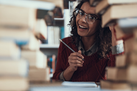 Positive pretty young lady doing homework with books in front of her and laughing while sitting with a pencil in her handの写真素材