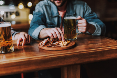 Close up of male hands with mugs of lager beer and junk food on wooden table. Gentleman in denim shirt on blurred backgroundの写真素材