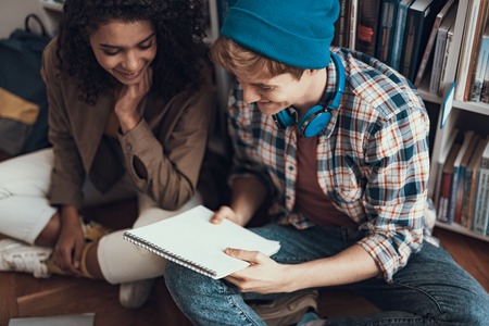 Cheerful happy young man in blue hat sitting with curly lady next to the bookcase and smiling while holding his notebookの写真素材
