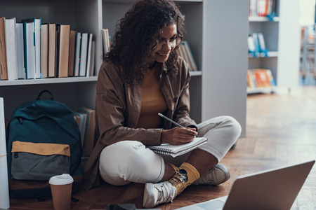 Positive confident student doing her homework on the floor of the library and smiling while making notes and looking at the screen of laptopの写真素材
