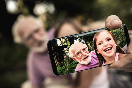 Senior gray-haired man in glasses with funny laughing girl shooting themselves on smartphone while sitting in summer garden. Focus on phone screenの写真素材