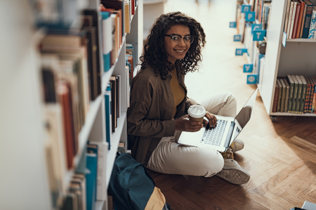 Emotional confident young lady sitting on the floor of the library and enjoying her coffee while holding modern laptopの写真素材