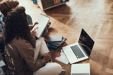 Busy students sitting against the bookcase on the floor and looking at the notes in their notebook while studyingの写真素材