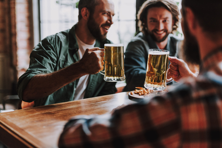 Handsome bearded men holding mugs of beer while sitting at the table and laughing. Focus on male hands with beveragesの写真素材
