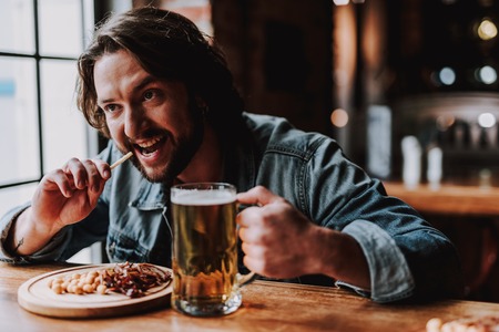 Portrait of attractive young gentleman eating appetizer and drinking beer while sitting at the tableの写真素材