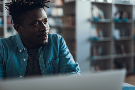 Close up of calm young man sitting in the library with bookshelves on the background and looking into the distance with a smileの写真素材