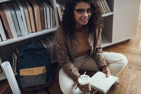 Pleased pretty curly student sitting on the floor with a cup of coffee and smiling while being alone...