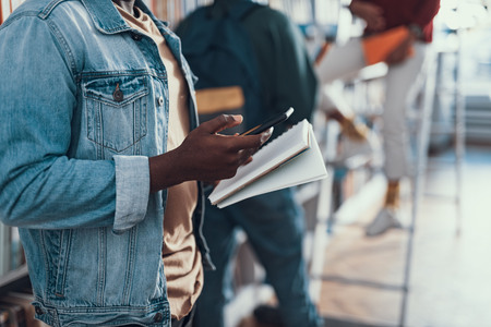 Afro-American student in casual clothes standing and holding a notebook in his left hand and a modern smartphone in his right handの写真素材