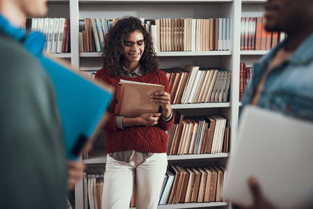 Positive relaxed young lady standing in the library with folders in her hands and smilingの写真素材