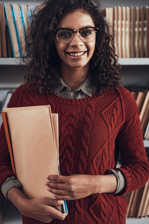 Glad relaxed student in warm sweater looking happy and smiling while standing near the bookcase and holding foldersの写真素材