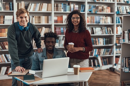 Positive emotional young Afro-American student sitting at the table with modern laptop while his two fellow students standing near and smilingの写真素材