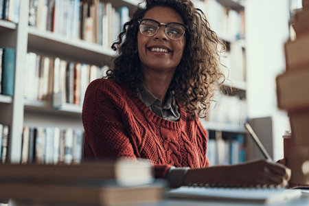 Positive emotional curly student in warm sweater holding a pencil and smiling while sitting in the library and looking into the distanceの写真素材