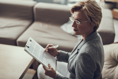 Calm experienced middle aged psychologist wearing glasses and looking away while holding clipboard with notesの写真素材