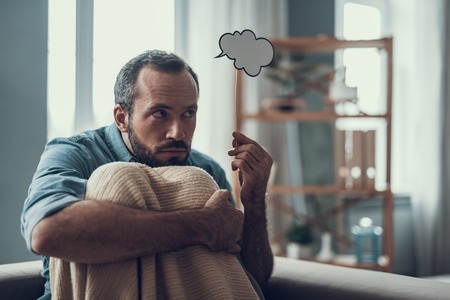 Depressed bearded middle aged man hugging his knees and looking away while holding a grey thought cloudの写真素材