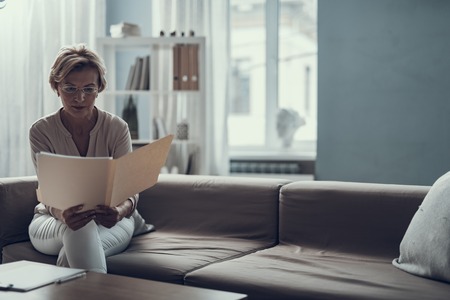 Thoughtful concentrated psychotherapist sitting on the sofa with a folder in her hands and looking attentively at the documents in itの写真素材