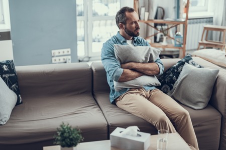Bearded middle aged man sitting on the sofa and frowning while holding a pillow and looking awayの写真素材