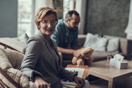 Cheerful psychotherapist wearing glasses and having notes on her laps while her client sitting on the background with teddy bearの写真素材