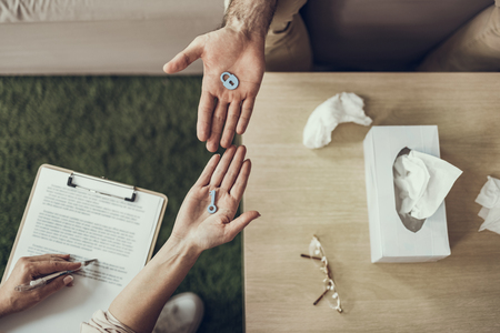 Psychotherapist with clipboard on the laps holding a little toy key while her client sitting opposite her and holding toy lockの写真素材