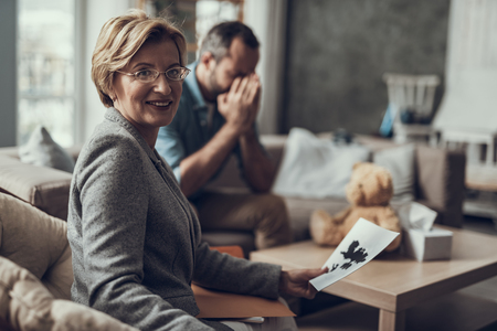 Positive professional psychotherapist sitting in the armchair with her client on the background and smiling while holding a picture from testの写真素材