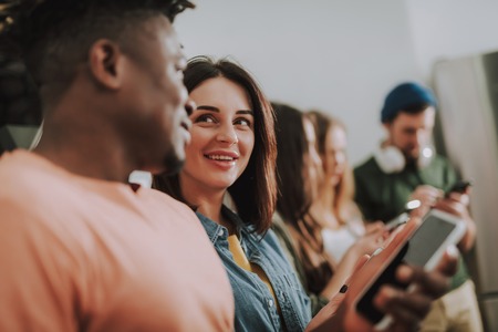 Portrait of beautiful girl gazing at friend and smiling while he holding smartphone.