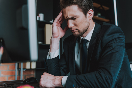 Unbearable headache. Close up of young man in elegant suit sitting at the table with his eyes closed and touching head while having sudden painの写真素材