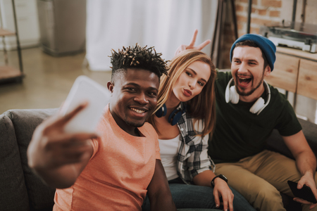 Portrait of handsome afro american guy holding smartphone and taking photo while his friends looking at phone camera and smilingの写真素材