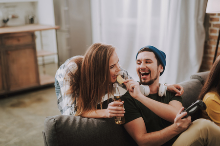 Taste this. Portrait of bearded guy in hat giving snacks to lady while she holding bottle of beerの写真素材